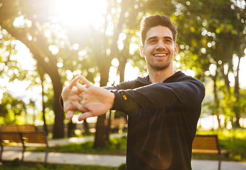 A man in a black sweatshirt stretches his arms in a park.