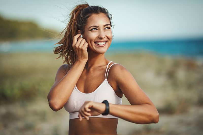 A woman in a sports bra smiles as she puts in her ear buds and checks her fitness tracker.