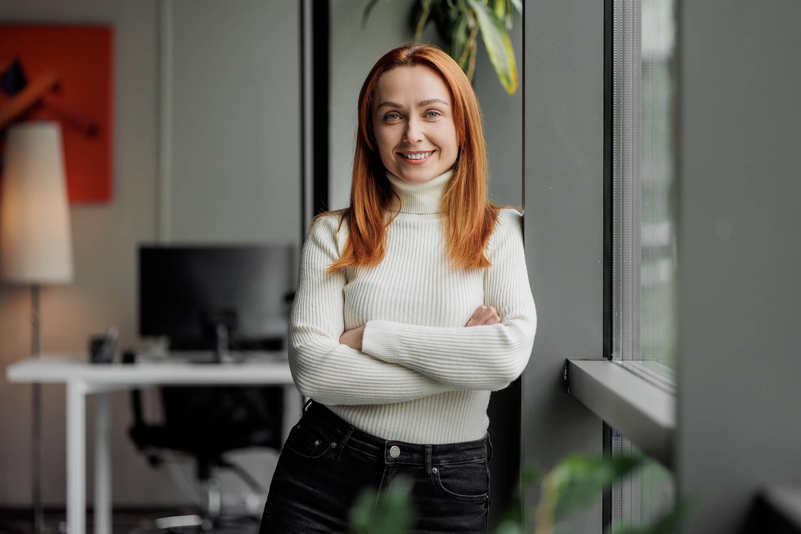 A woman with red hair smiles at the camera while standing by a window in an office.